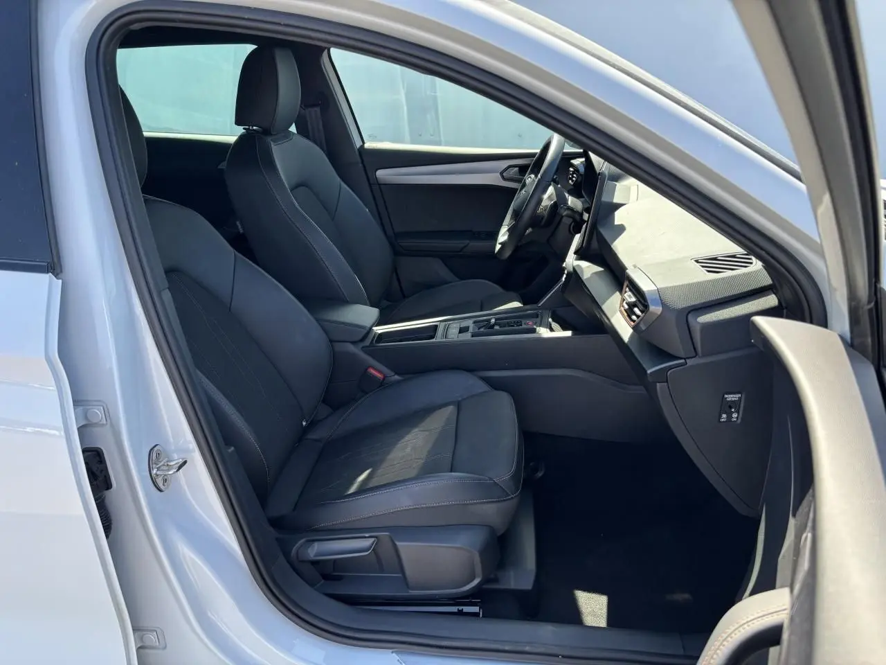 Vue intérieure côté conducteur d'une CUPRA Leon Sportstourer blanche Nevada, sièges noirs et tableau de bord moderne.