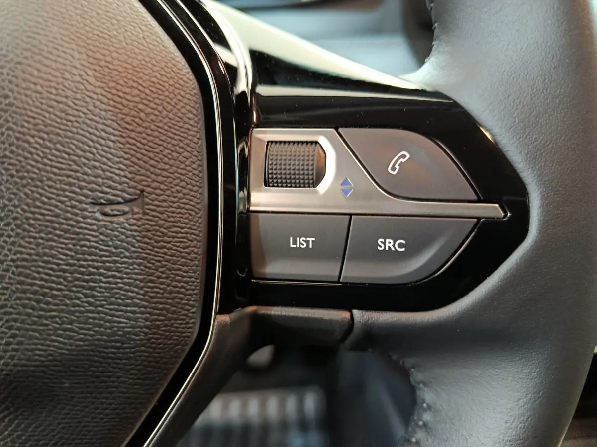 Gros plan sur les commandes gauche du volant noir du Peugeot 208 PureTech 100 S&S Allure gris clair, avec boutons LIST et SRC.