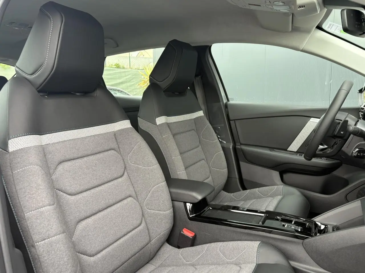 Vue intérieure côté conducteur de la Citroën C4 2024, sièges tissu gris avec accoudoir central noir brillant.