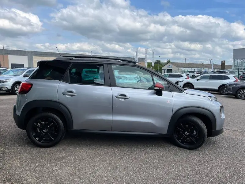 Profil droit d'un Citroën C3 Aircross gris acier avec toit noir et rétroviseur rouge sur parking extérieur.