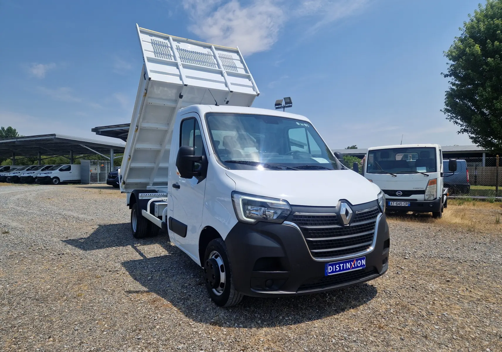 Renault Master Benne blanc vu 3/4 avant droit, benne relevée et roues jumelées visibles sur terrain gravillonné.