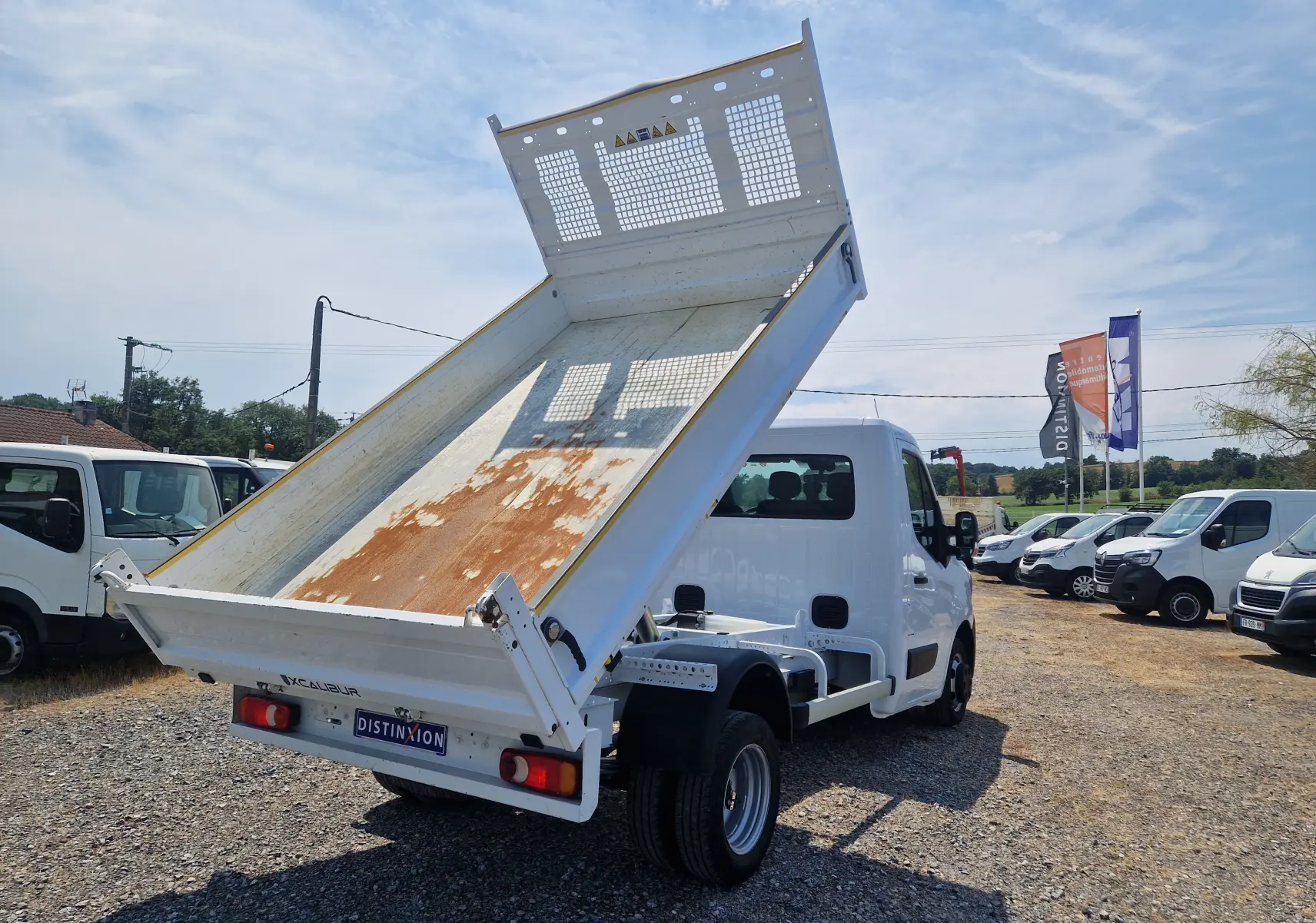 Vue 3/4 arrière droite d’un Renault Master blanc avec benne basculante relevée et roues jumelées, sur un terrain extérieur.