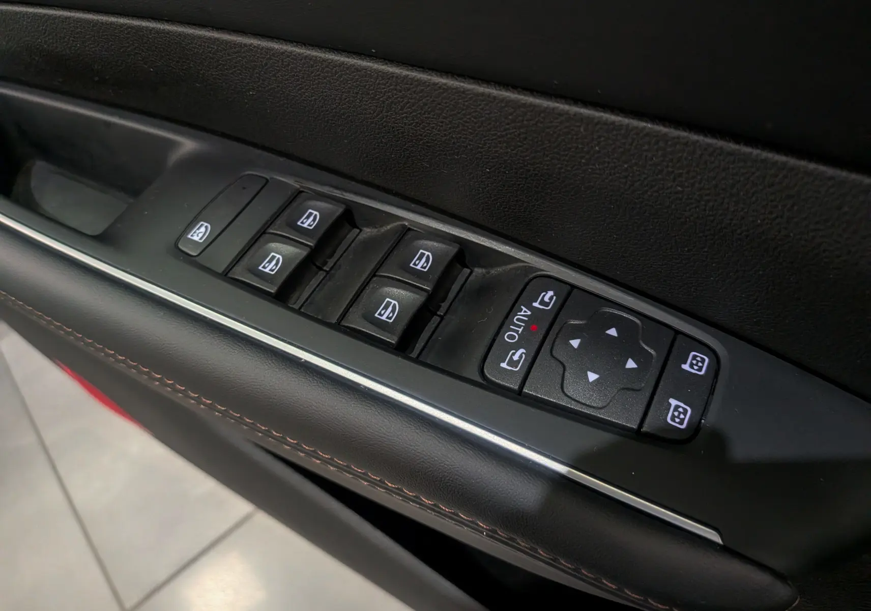 Gros plan sur les commandes des vitres et rétroviseurs électriques côté conducteur en cuir noir avec surpiqûres rouges.