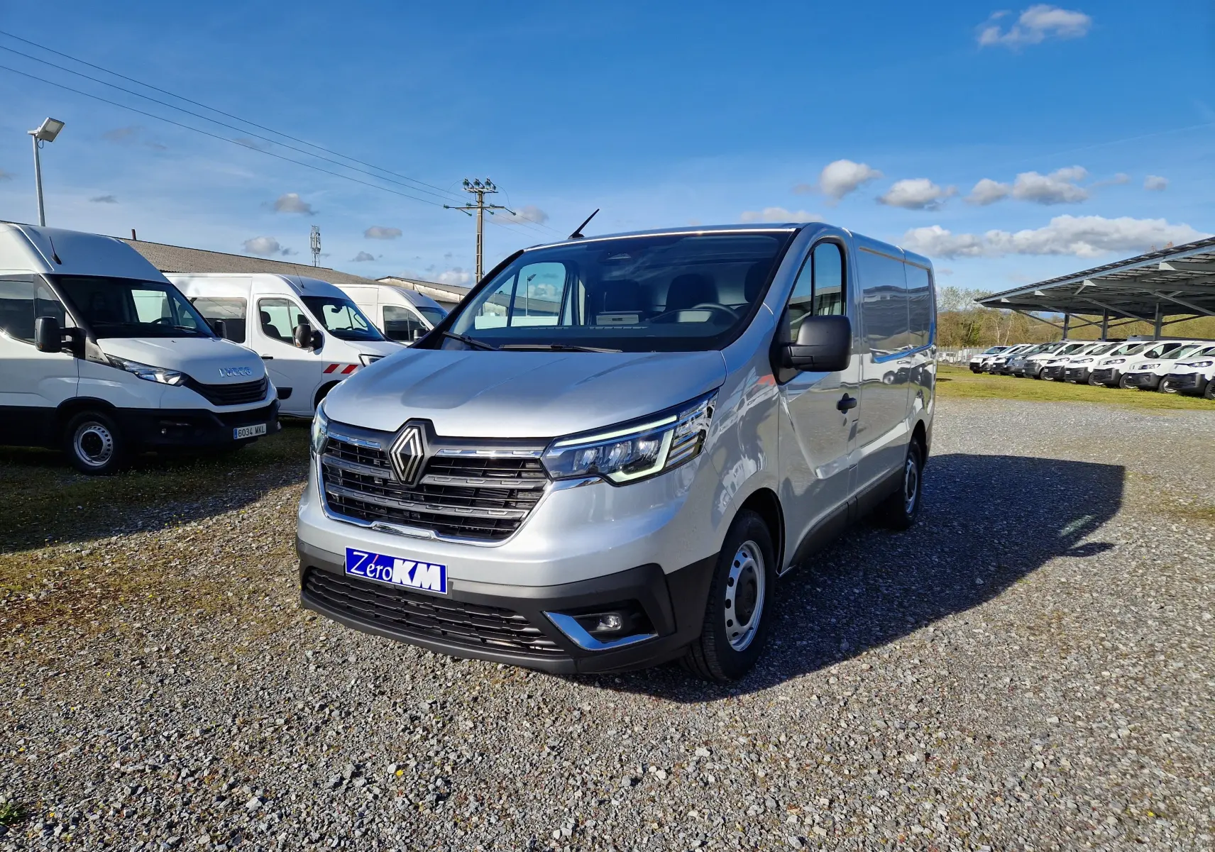 Renault Trafic Fourgon gris Highland vu en 3/4 avant droit sur parking avec plusieurs autres utilitaires blancs.