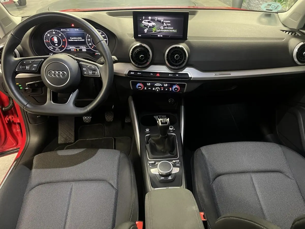Vue intérieure avant de l’Audi Q2 rouge 2024, volant sport cuir, écran central et boîte manuelle visible.