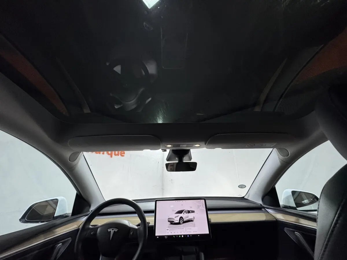 Vue intérieure frontale du tableau de bord et écran tactile central d'une Tesla Model Y blanche avec toit panoramique.