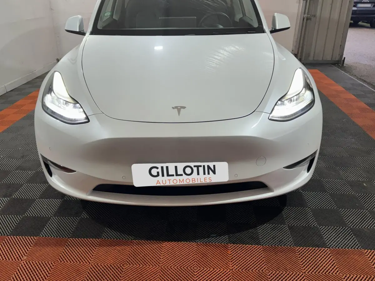 Vue frontale d'une Tesla Model Y blanche avec phares LED allumés sur sol à damier noir et orange.