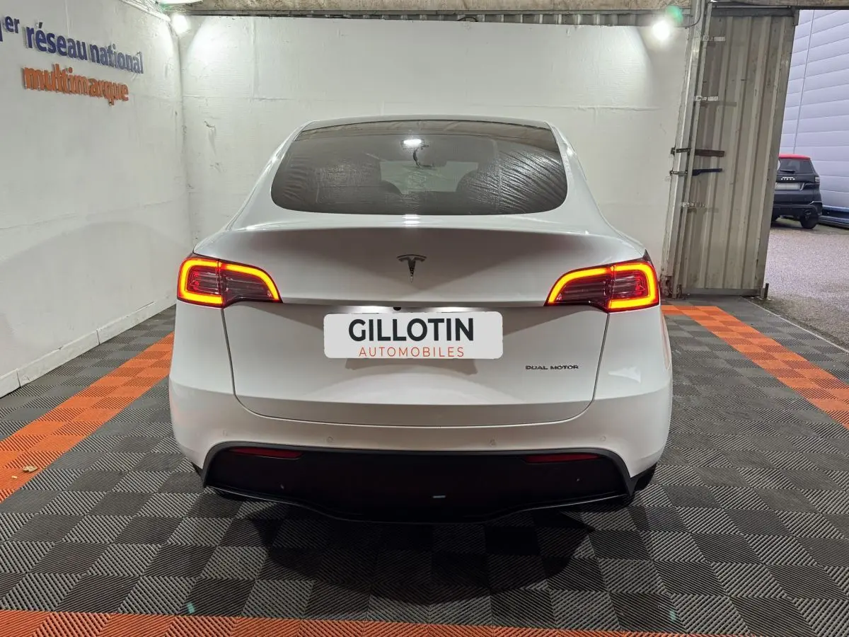 Vue arrière d'une Tesla Model Y blanche avec feux arrière allumés dans un garage éclairé.