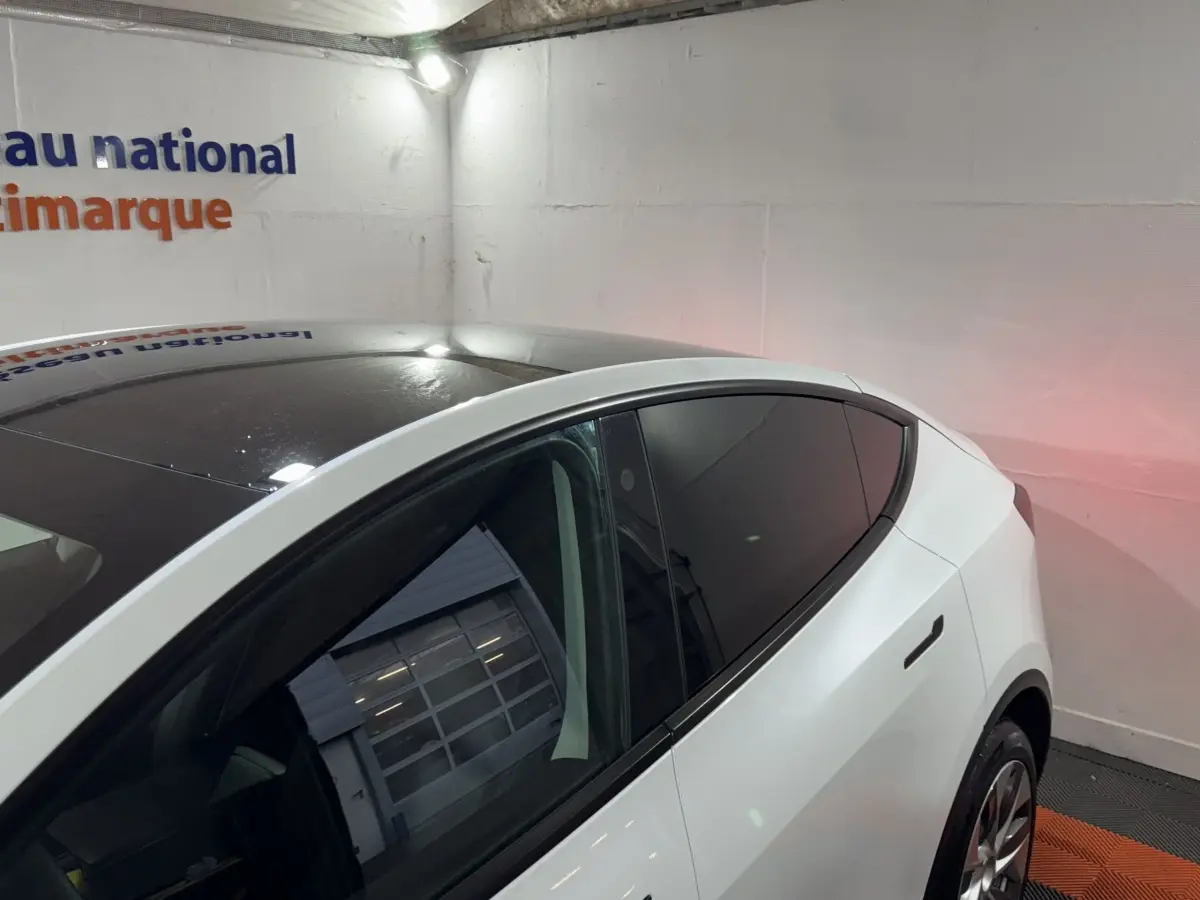 Vue 3/4 arrière droite d'une Tesla Model Y blanche avec toit panoramique noir et vitres teintées dans un garage.