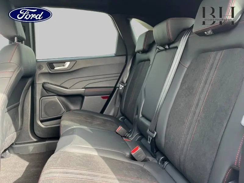 Vue intérieure arrière côté gauche du Ford Kuga 2026, sièges en suédine noire avec surpiqûres rouges et ceintures grises.