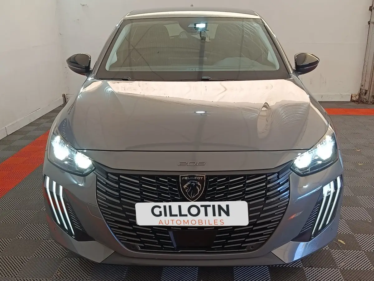 Vue avant d'une Peugeot 208 gris foncé avec phares allumés et calandre noire distinctive en intérieur showroom.