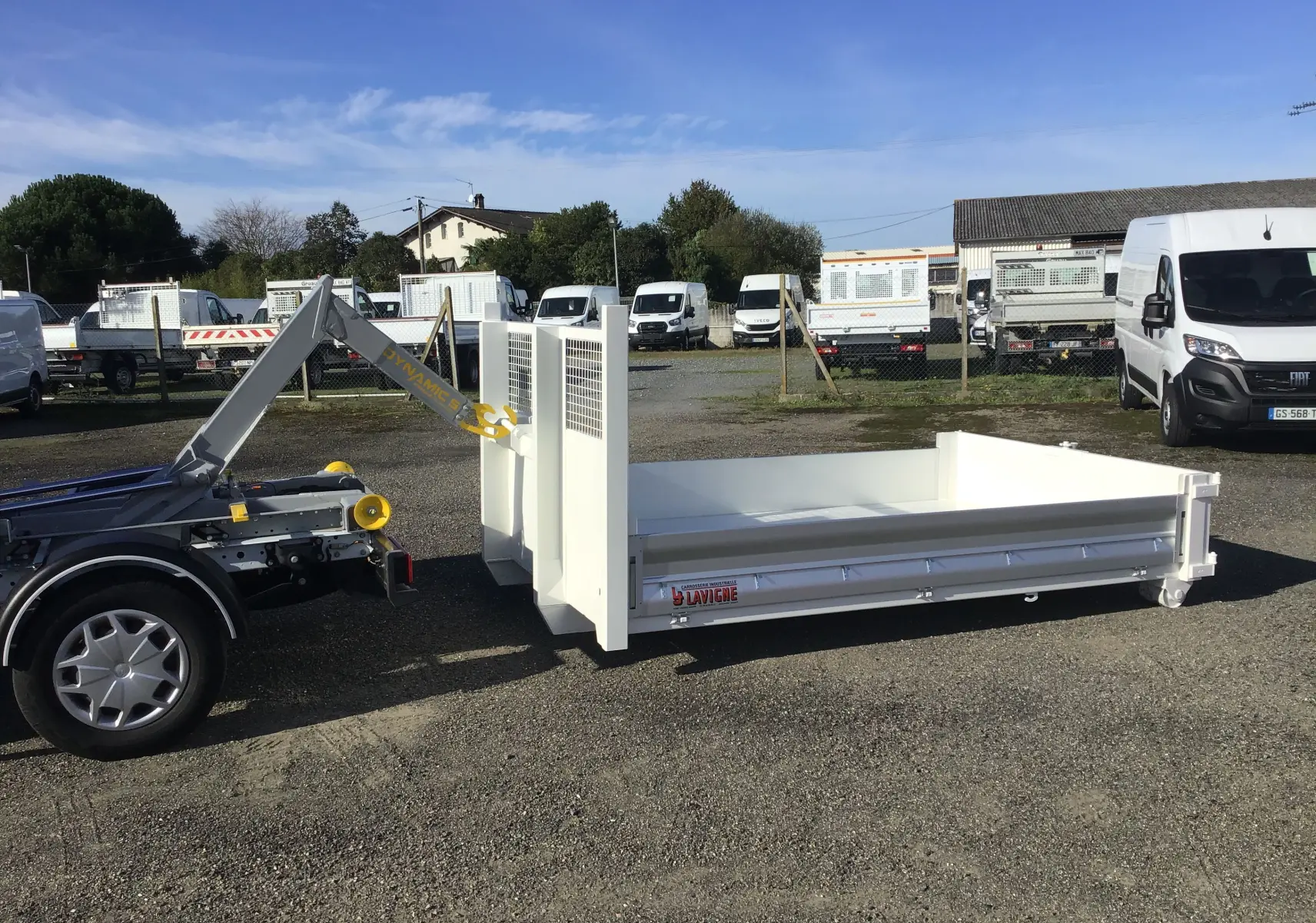 Vue latérale gauche du châssis cabine Ford Transit blanc avec bras de levage et benne polybenne détachée au sol.