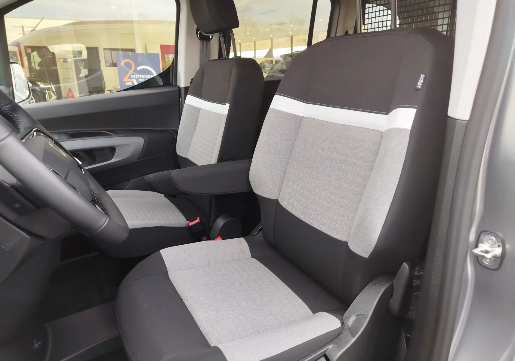 Vue intérieure côté conducteur du Citroën Berlingo 2025 gris acier, sièges tissu bicolore avec accoudoir central.