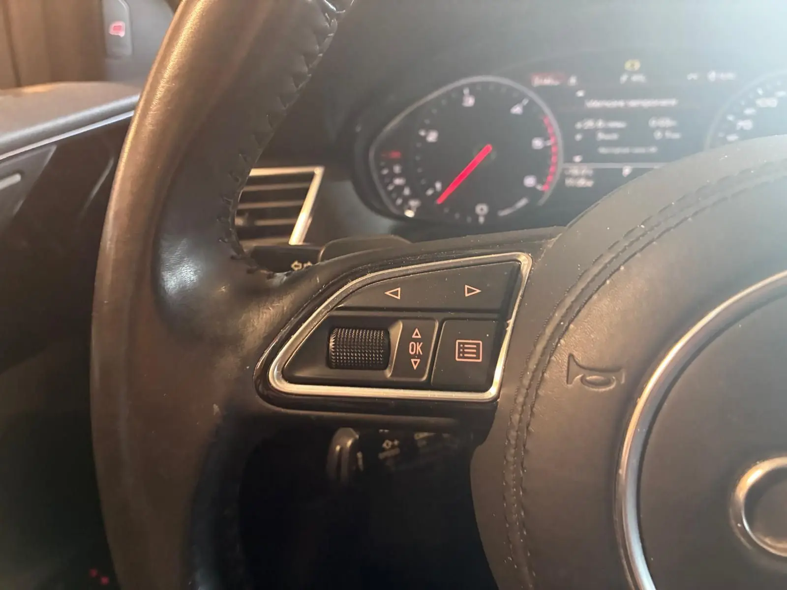Gros plan sur le côté gauche du volant en cuir noir de l’Audi A8 2016 avec commandes intégrées et tableau de bord flou en arrière-plan.