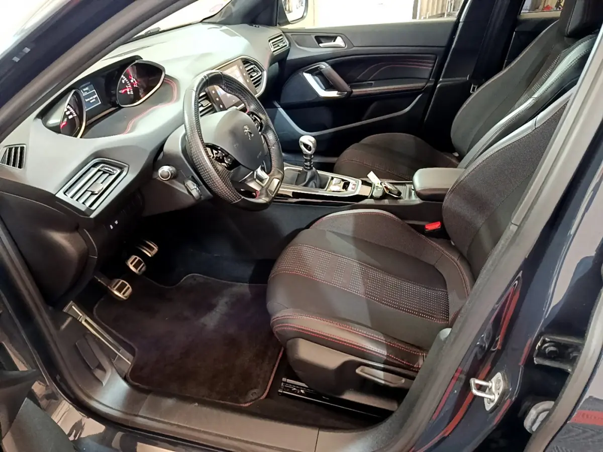 Intérieur avant droit de Peugeot 308 gris foncé, volant cuir, sièges tissu noirs avec surpiqûres rouges, boîte manuelle visible.