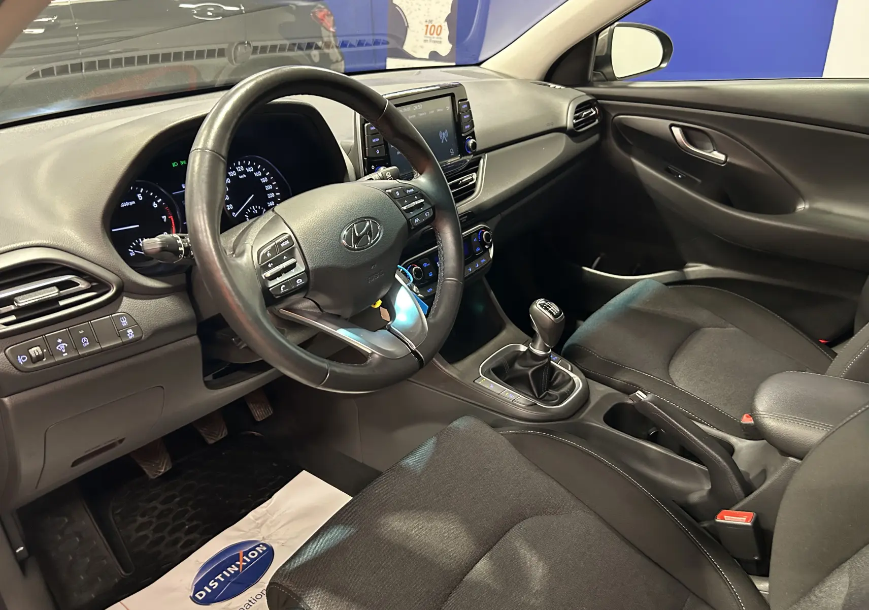 Vue intérieure de la Hyundai i30 SW 2022 côté conducteur, montrant le volant cuir, la boîte manuelle et les sièges tissu noirs.