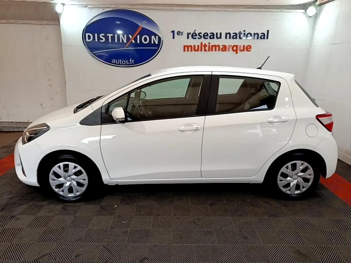 Profil côté gauche d'une Toyota Yaris blanche 2019, compacte, avec jantes alliage et arrière arrondi, en intérieur showroom.