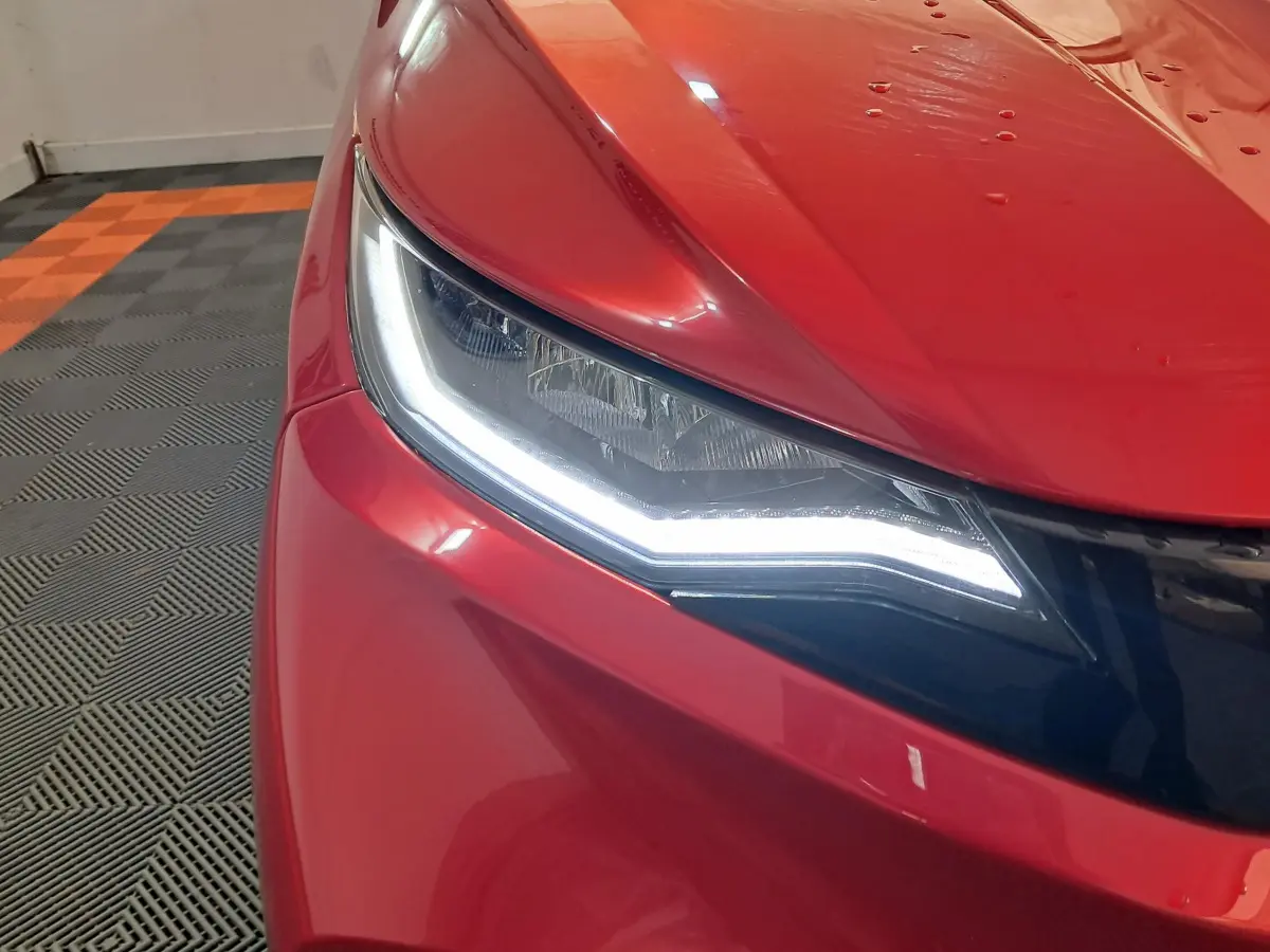 Gros plan sur le phare avant gauche allumé d'une Aixam Ambition rouge 2025 dans un garage au sol carrelé.