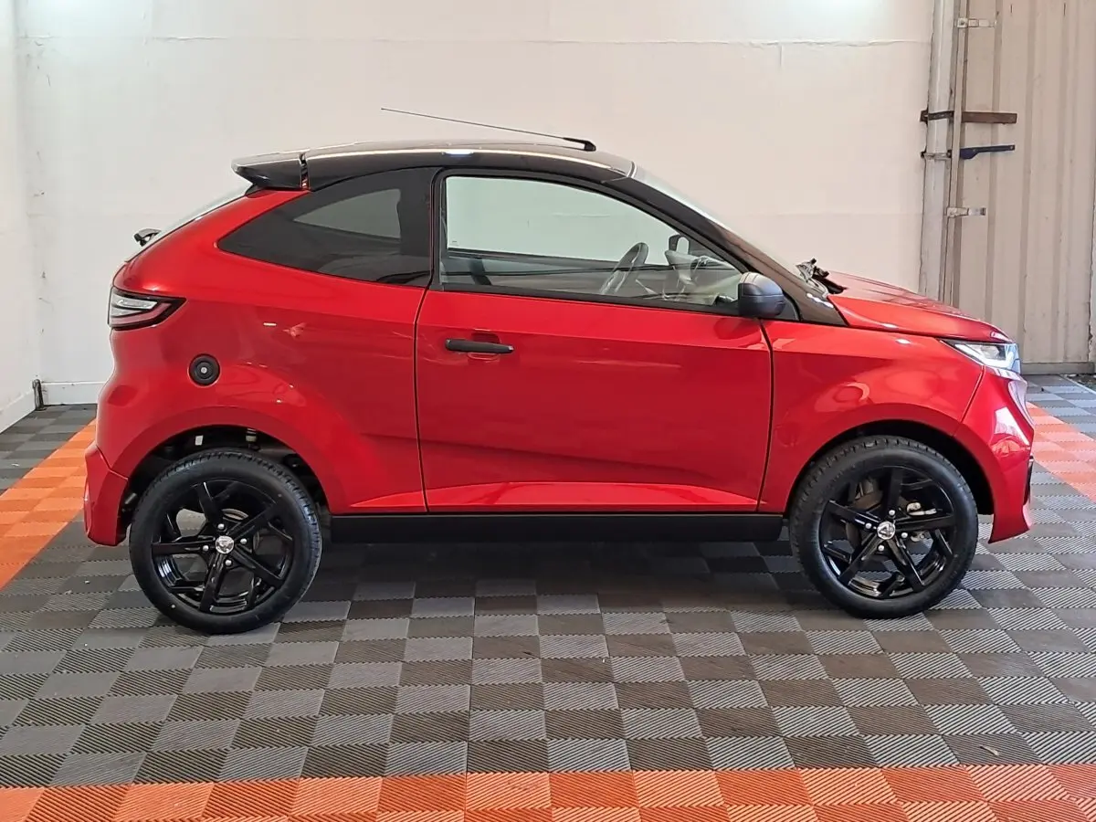 Profil côté gauche de l'AIXAM AMBITION rouge 2025 avec toit noir et jantes noires sportives dans un intérieur lumineux.