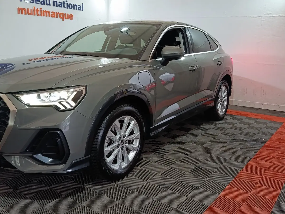 Audi Q3 Sportback gris foncé en 3/4 avant droit, phare allumé et jantes alliage visibles dans un showroom.
