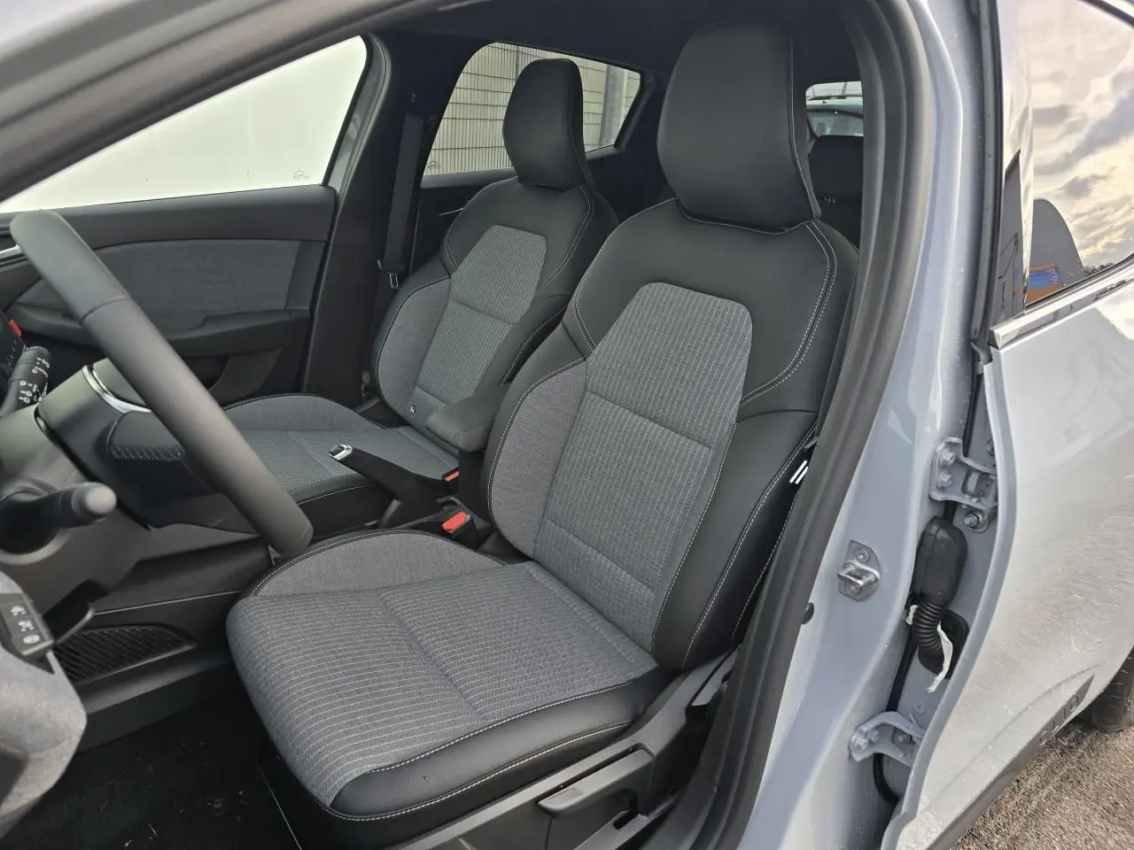 Vue intérieure côté conducteur de la Renault Clio gris Rafale, montrant les sièges bi-matière gris et noir avec surpiqûres blanches.