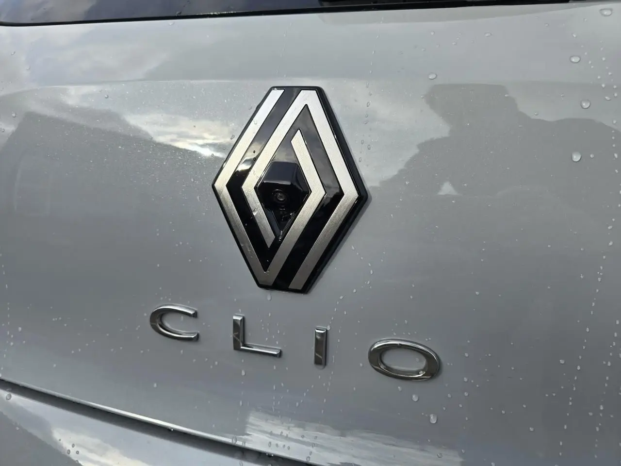 Gros plan sur le logo Renault et le badge Clio sur la malle arrière gris rafale métal avec gouttes d'eau visibles.