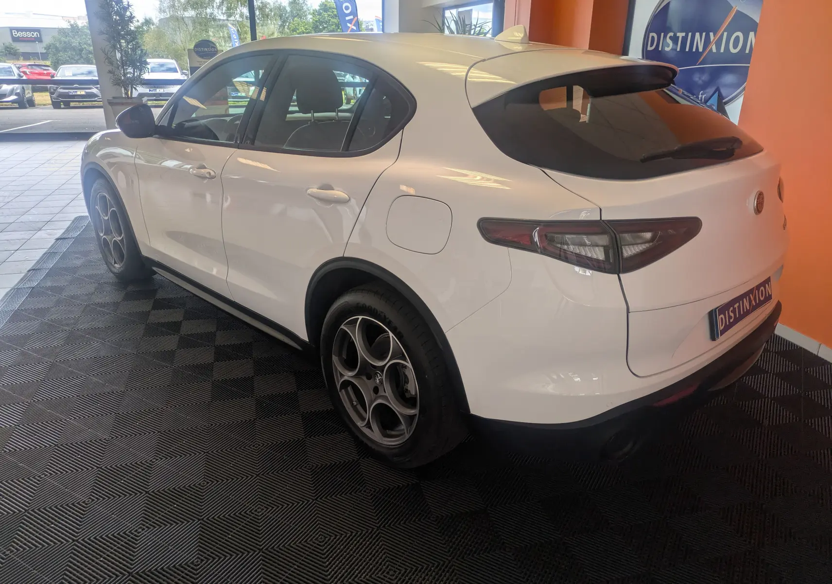 Vue 3/4 arrière droite d'un Alfa Romeo Stelvio blanc 2023 dans un showroom avec jantes bi-ton et vitres teintées.