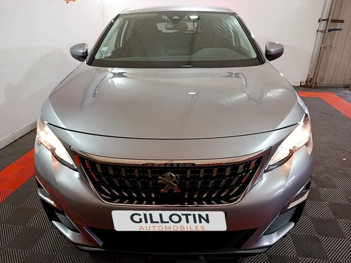 Vue avant d’un Peugeot 3008 gris clair avec calandre noire et phares allumés dans un garage.