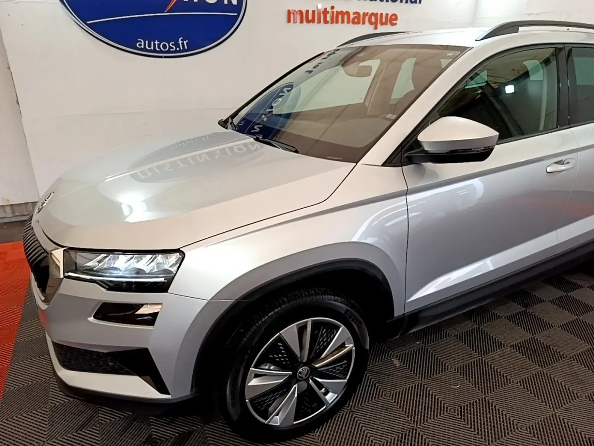 Vue 3/4 avant droit d'un Skoda Karoq gris clair avec jantes alliage et phares LED allumés en intérieur showroom.