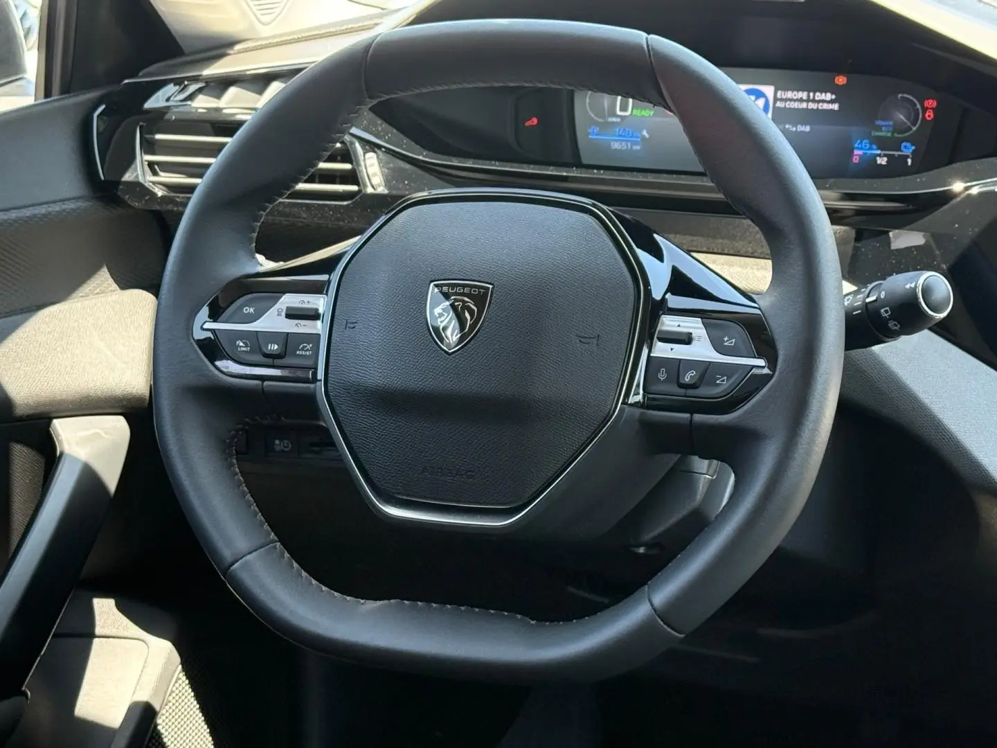 Vue rapprochée du volant noir multifonction de la Peugeot e-308 2025 avec tableau de bord digital en arrière-plan.