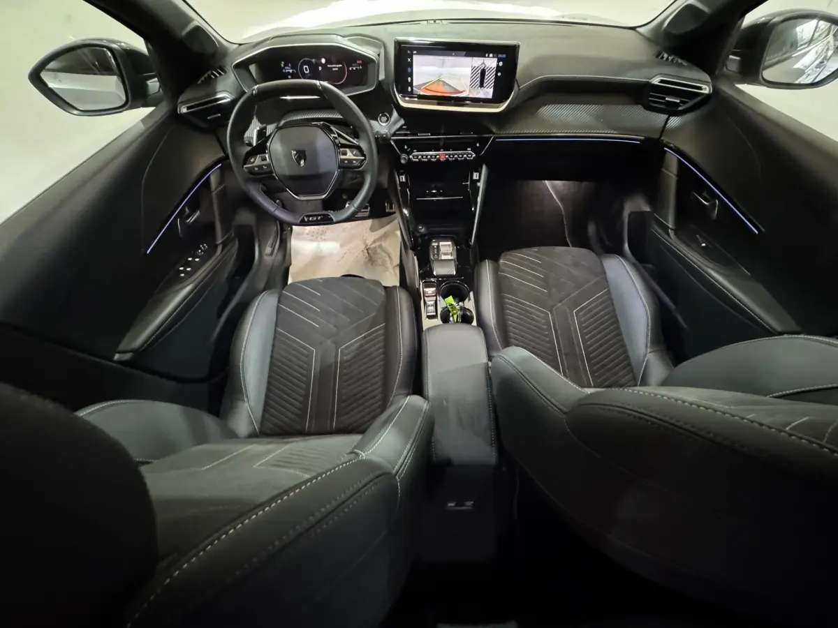 Vue intérieure avant de la Peugeot 208 Hybrid GT 2025, tableau de bord noir avec écran tactile et éclairage d'ambiance bleu.
