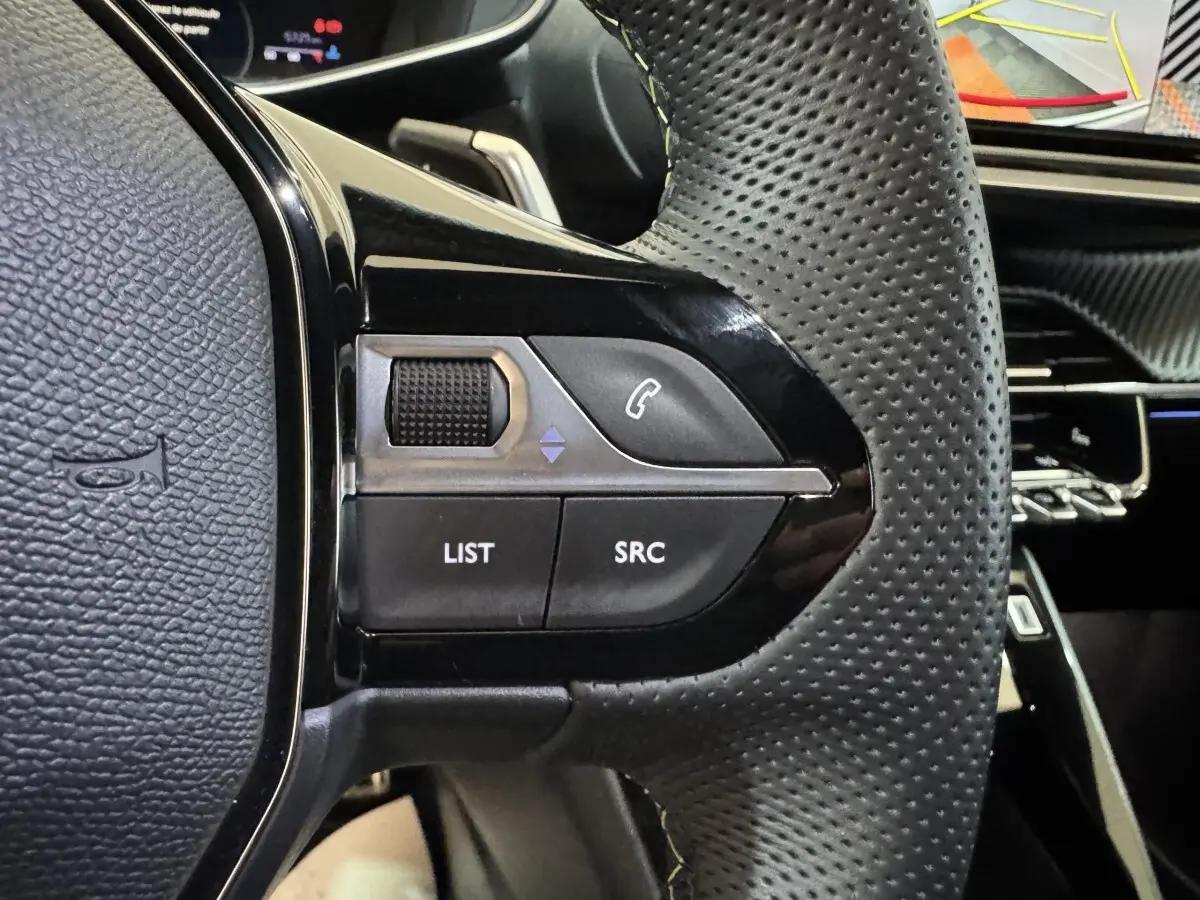 Gros plan sur les commandes gauche du volant en cuir perforé noir du Peugeot 208 Hybrid gris clair, avec tableau de bord en arrière-plan.