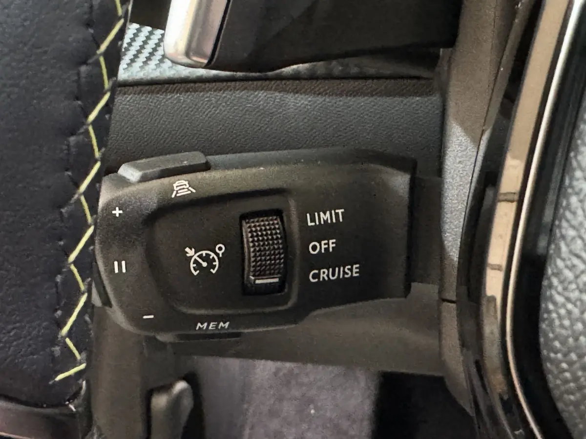 Gros plan sur la commande de régulateur de vitesse au volant cuir noir avec surpiqûres jaunes d’une Peugeot 208 Hybrid 136 eDCS6 GT gris clair.