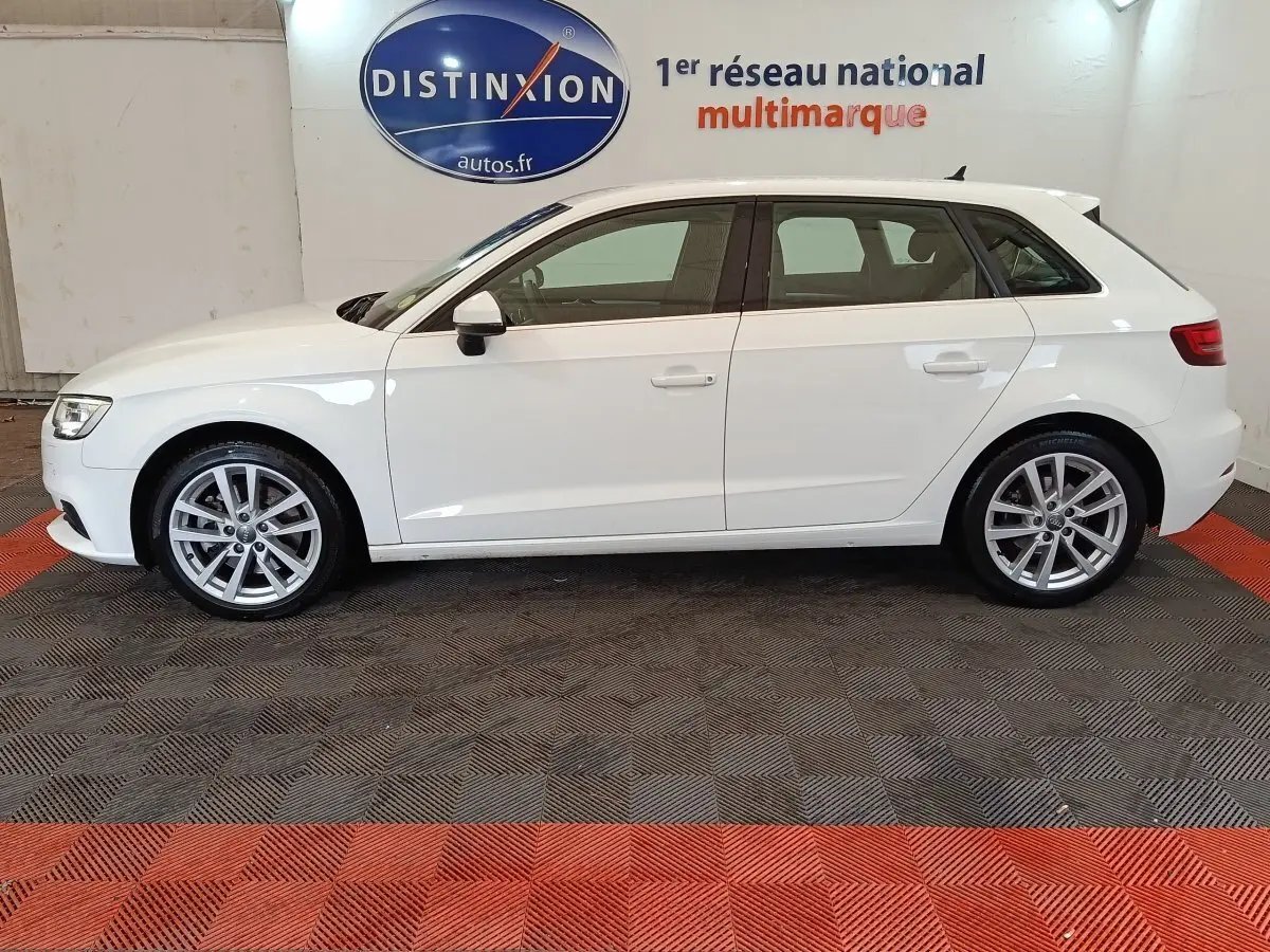 Profil latéral droit d'une Audi A3 blanche 35 TDI 150 S tronic Business Line sur sol noir et rouge en intérieur showroom.