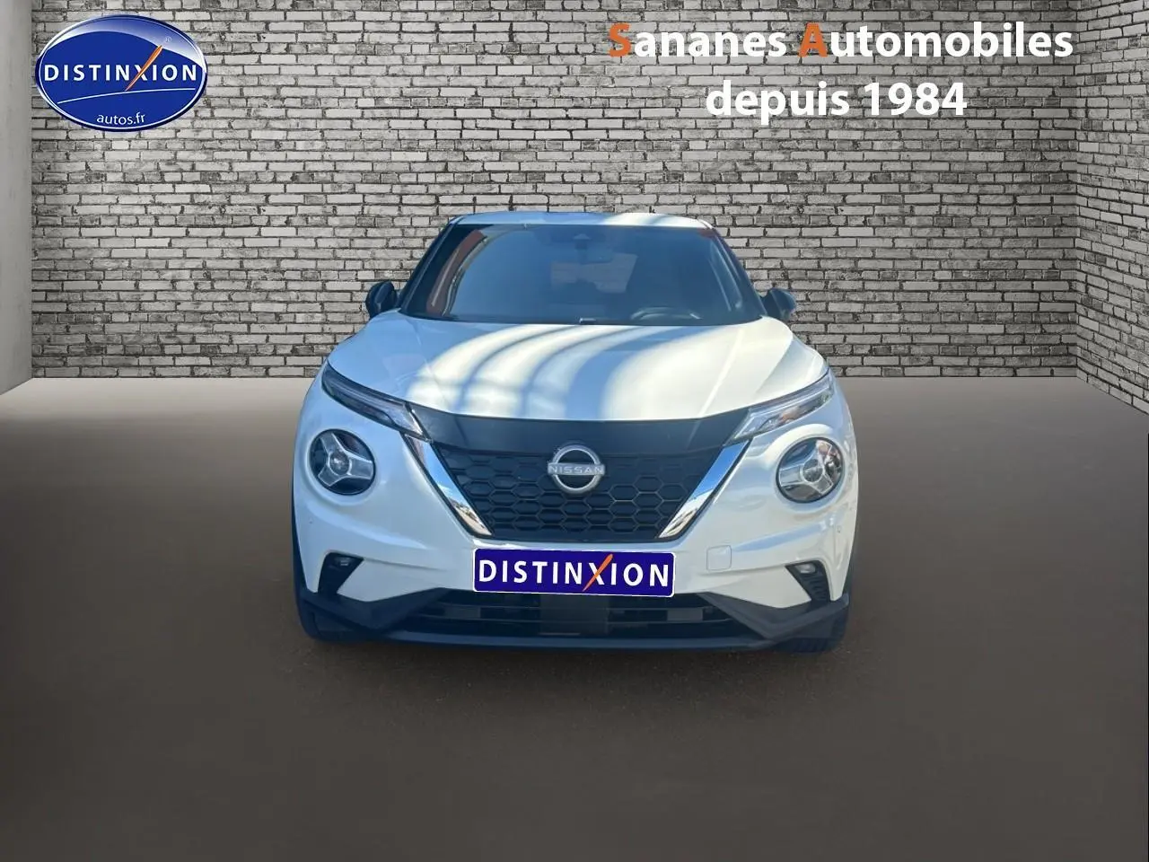 Vue frontale d'un Nissan Juke blanc 2024 avec calandre noire et phares ronds distinctifs sur fond de mur en briques.