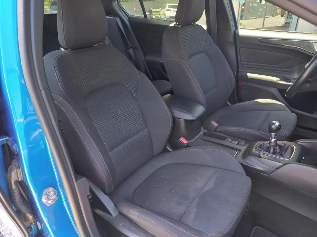 Vue intérieure côté conducteur d'une Ford Focus bleu 2019, sièges noirs avec surpiqûres rouges et levier de vitesse manuel.