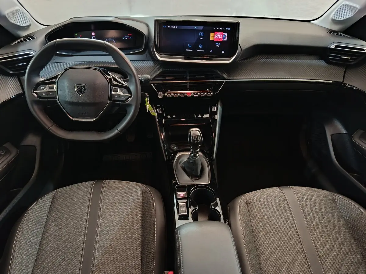 Vue intérieure avant de la Peugeot 208 PureTech 100 S&S Allure 2025, tableau de bord noir avec écran tactile et boîte manuelle.