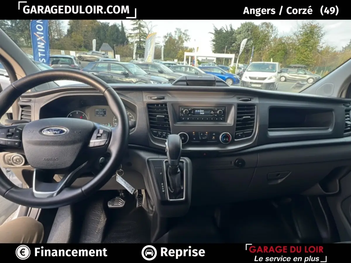 Vue intérieure avant du Ford Transit Custom blanc 2023, montrant le volant cuir multifonctions et la console centrale avec boîte auto.