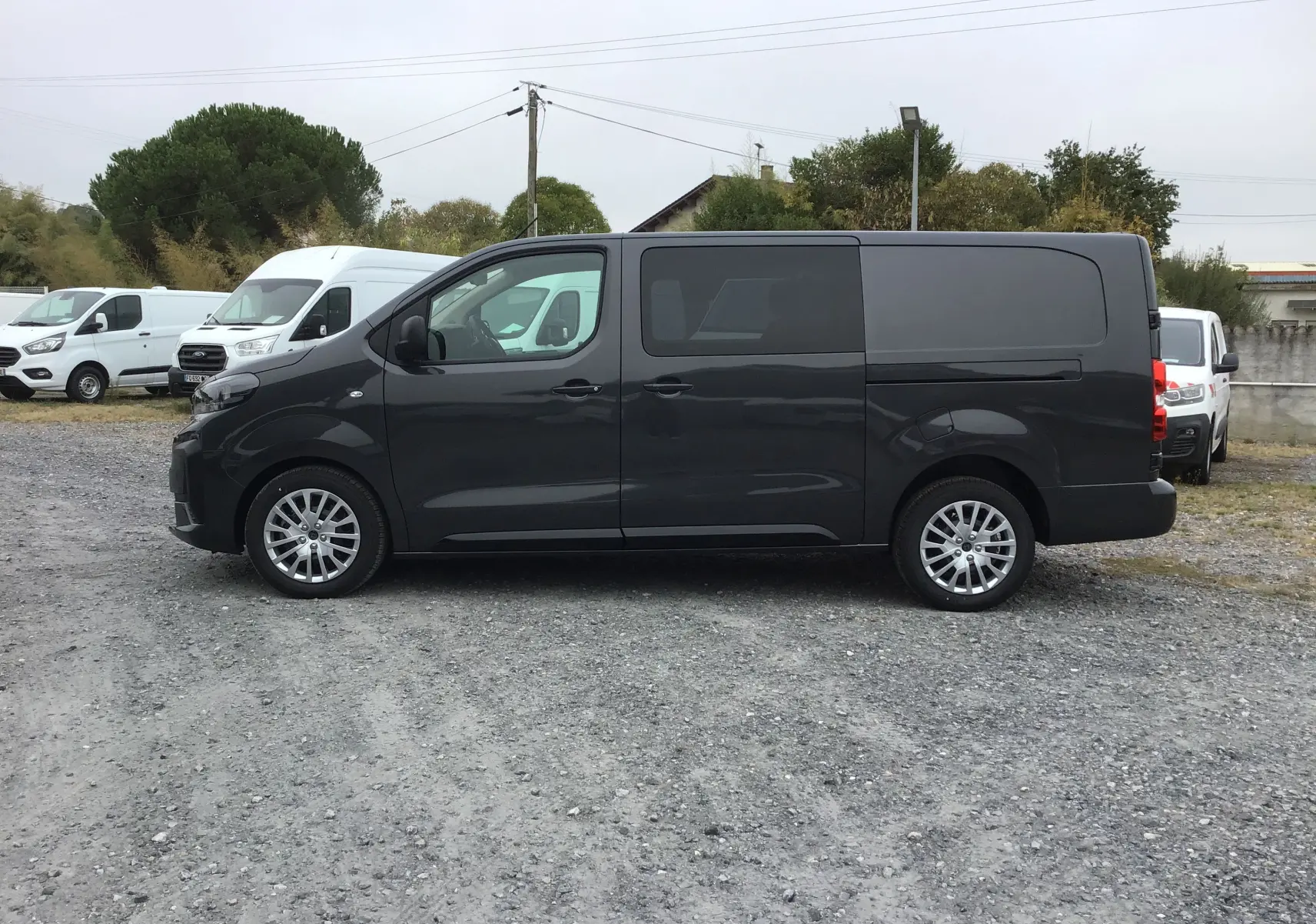 Peugeot Expert Cabine Approfondie gris titane vu de profil côté gauche sur un sol gravillonné, avec plusieurs véhicules blancs en arrière-plan.