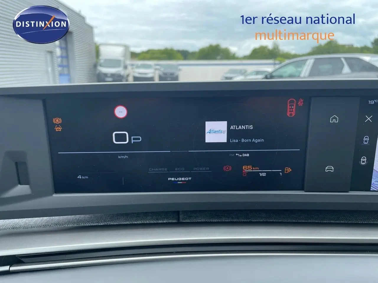 Tableau de bord numérique du Peugeot 5008 Hybrid 2025 affichant la radio et l'autonomie, avec vue sur parking extérieur flou.