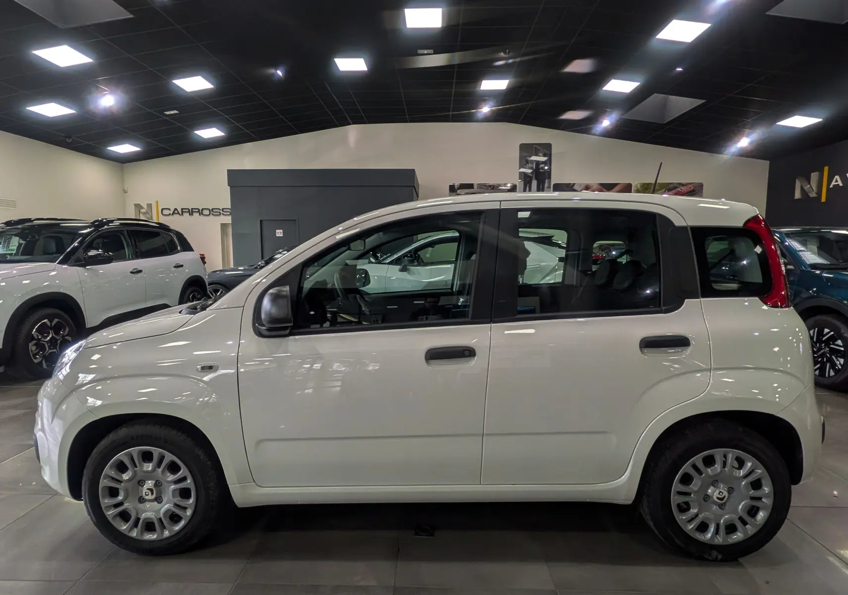 Vue de profil côté gauche d'une Fiat Panda 1.0 70 CH Hybrid City blanche Gelato en intérieur, avec jantes acier et rétroviseurs noirs.