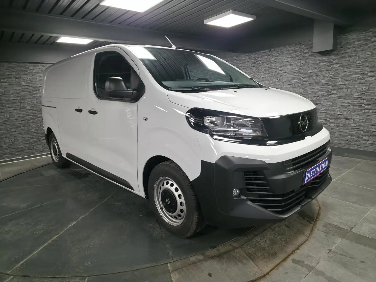 Opel Vivaro blanc en 3/4 avant droit, avec calandre noire et pare-chocs avant noir mat dans un intérieur gris.