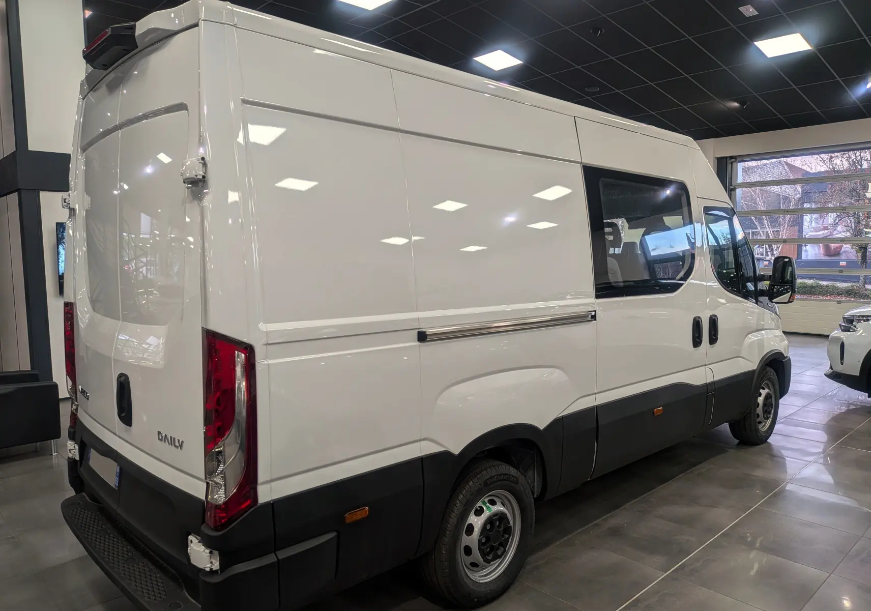 Vue 3/4 arrière droite d’un Iveco Daily Fourgon blanc 2025 avec porte latérale vitrée et pare-chocs noir.