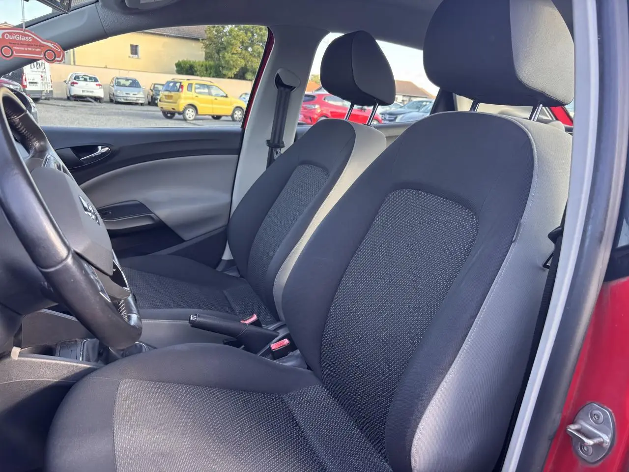 Vue intérieure côté conducteur de la SEAT Ibiza rouge clair 2016, montrant les sièges avant noirs et la portière ouverte.