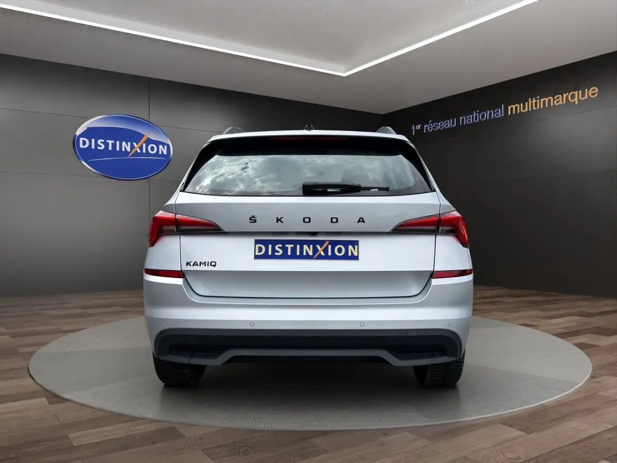 Vue 3/4 arrière droite d'un Skoda Kamiq gris clair avec jantes bi-ton et caméra de recul en showroom.