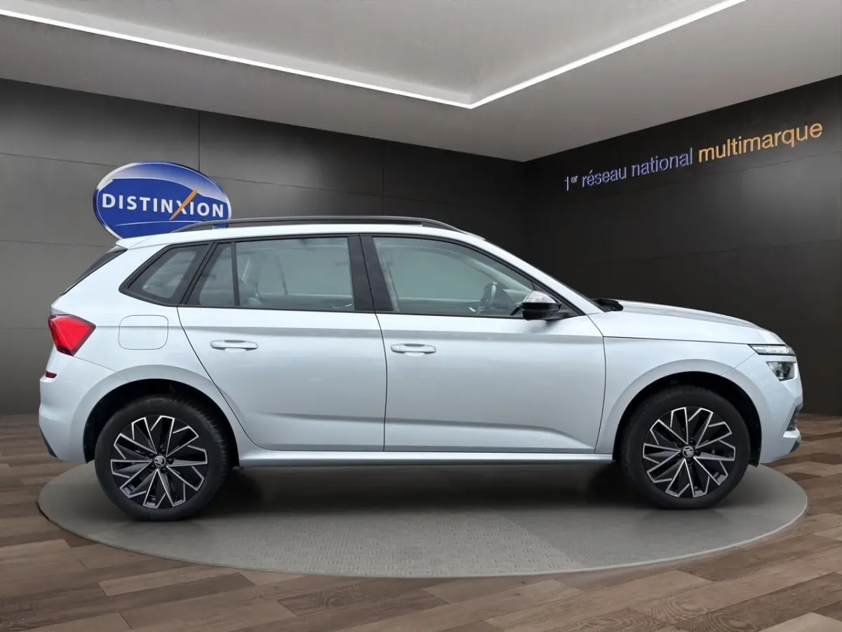 Vue 3/4 arrière droite d’un Skoda Kamiq gris clair avec jantes bi-ton et barres de toit noires.