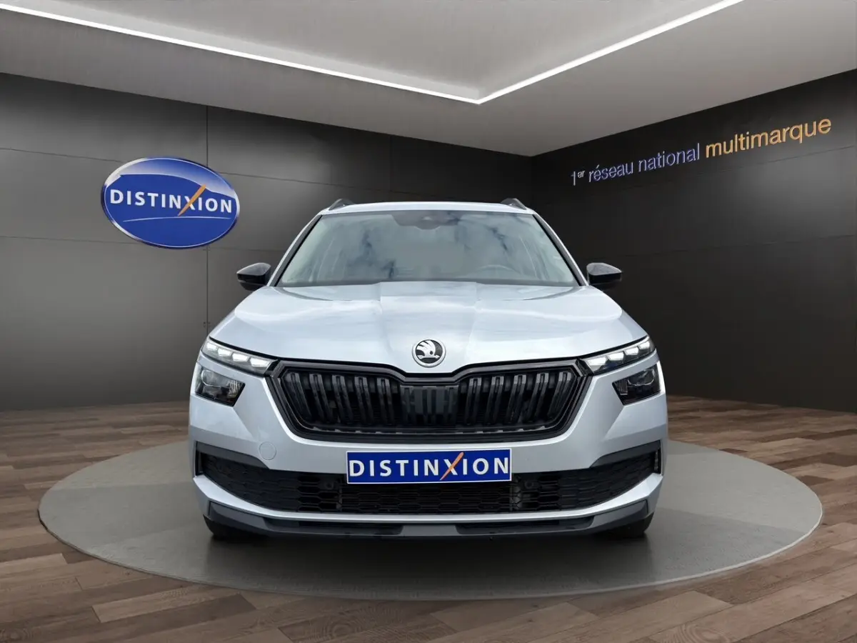 Vue 3/4 avant droite d'un Skoda Kamiq gris clair avec jantes bi-ton et rétroviseurs noirs dans un showroom.