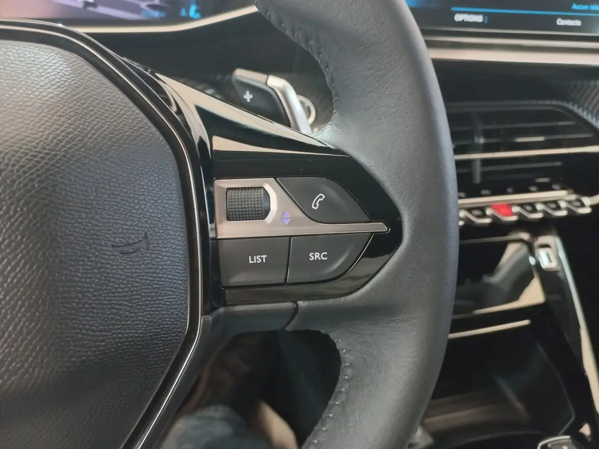 Gros plan sur le volant cuir noir de la Peugeot 208 2022 avec commandes multimédia et tableau de bord tactile en arrière-plan.