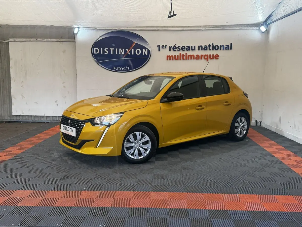 Peugeot 208 jaune vue 3/4 avant droit, avec calandre noire et feux LED, stationnée en intérieur sur sol damier noir et orange.