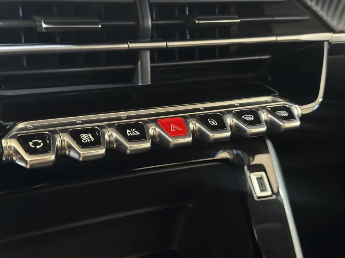 Gros plan sur la rangée de boutons métalliques du tableau de bord de la Peugeot 208 jaune, avec bouton rouge d’alerte au centre.